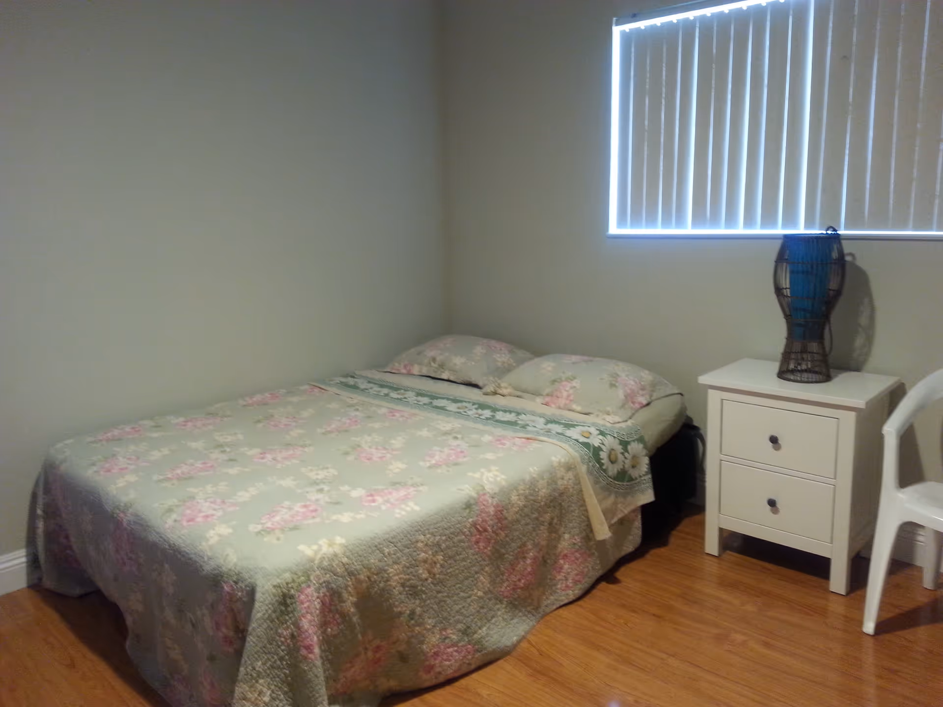 A simple bedroom with a bed covered in a floral bedspread, two pillows, a white nightstand with two drawers, a decorative lamp on the nightstand, a white plastic chair, and a window with vertical blinds.