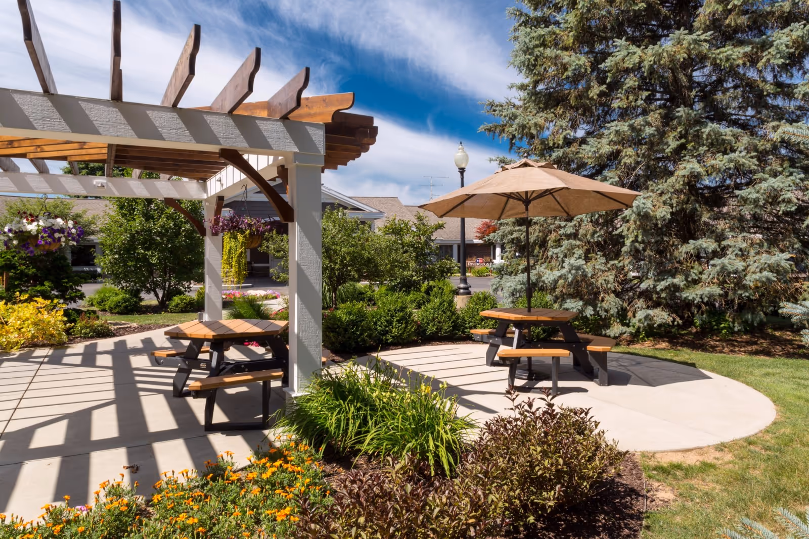 Outdoor patio area with wooden picnic tables, one with a large beige umbrella, surrounded by greenery, flowers, and trees under a blue sky with some clouds. A wooden pergola casts shadows on the concrete floor.