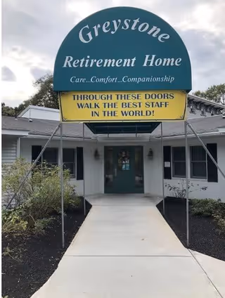 Entrance to Greystone Retirement Home with a green and yellow sign above the walkway that reads 'Greystone Retirement Home Care...Comfort...Companionship' and 'Through these doors walk the best staff in the world!'. The building is white with a gray roof and has a concrete pathway leading to the door, surrounded by some plants and bushes.