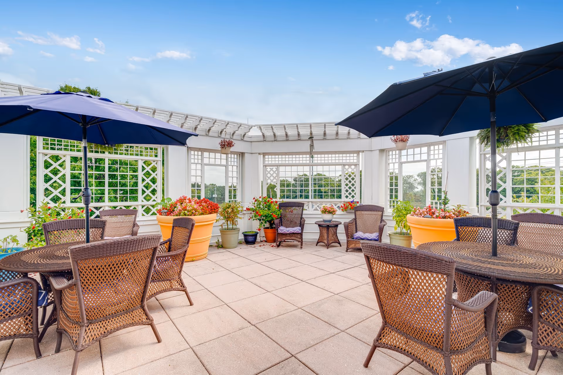 Outdoor patio area with wicker chairs and tables under large blue umbrellas, surrounded by white lattice fencing and large potted plants with flowers, under a partly cloudy sky.