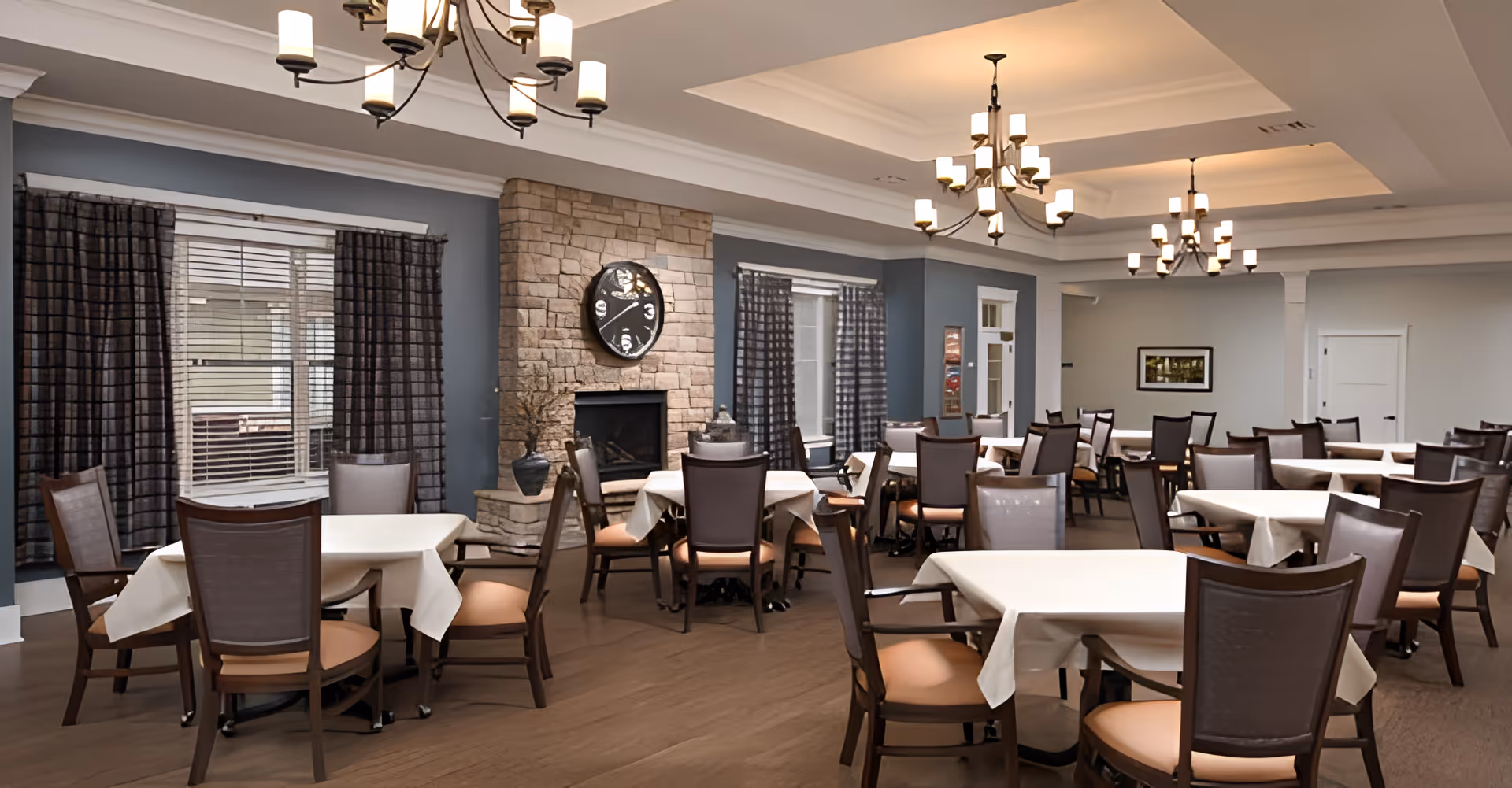 A spacious dining room with multiple tables covered in white tablecloths and surrounded by wooden chairs with cushioned seats. The room features a stone fireplace with a large clock above it, windows with dark curtains, and elegant chandeliers hanging from the ceiling. The walls are painted in neutral tones, creating a warm and inviting atmosphere.