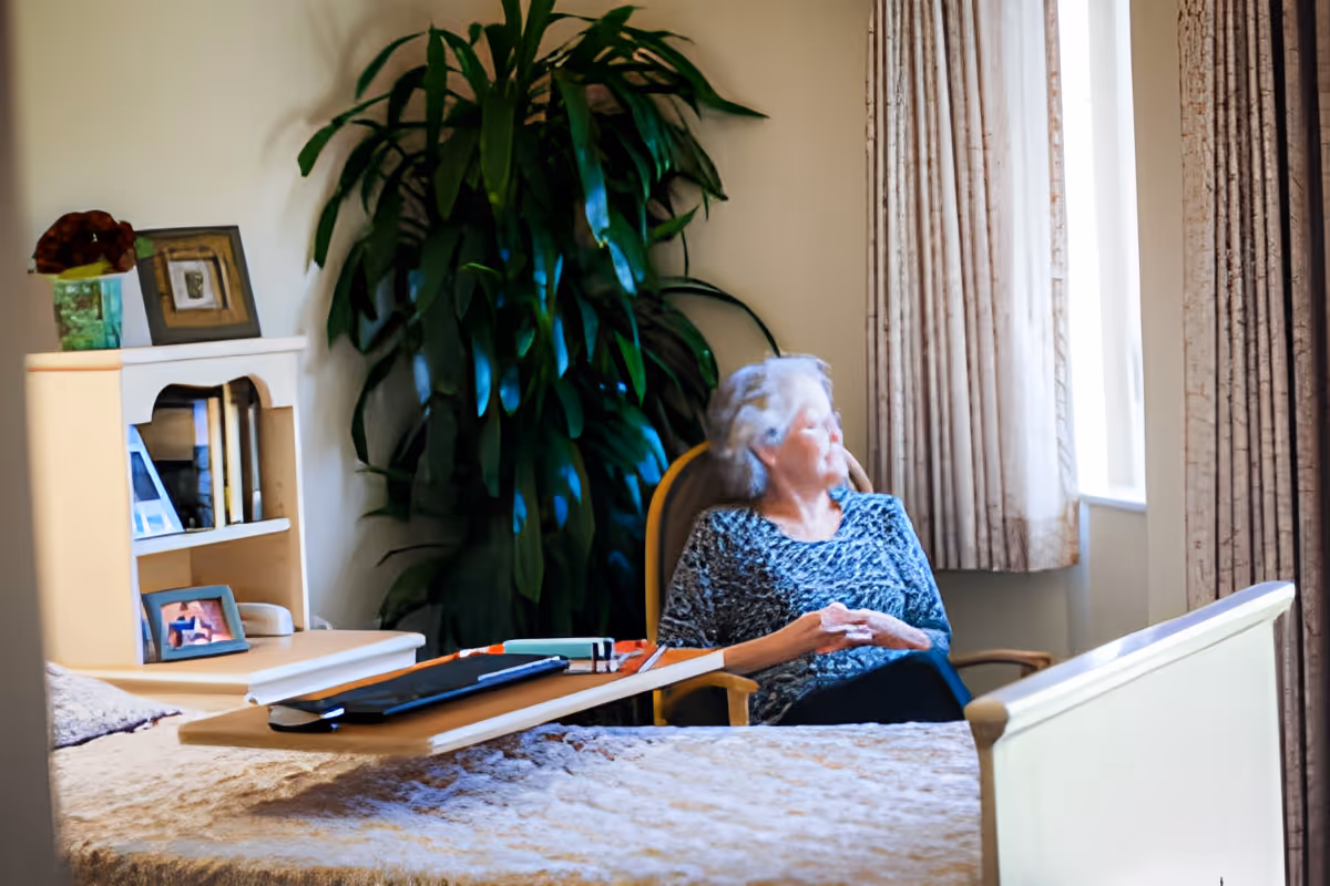 An elderly woman sits in a chair by a window in a nursing facility room with a bed, bedside table, and a large potted plant.