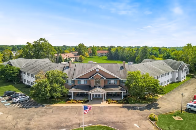 Aerial view of Emeritus at Canton senior living facility showing a large two-story building with a central entrance, surrounded by trees and greenery. There is a parking lot with several cars and an American flag in front of the building under a clear blue sky.