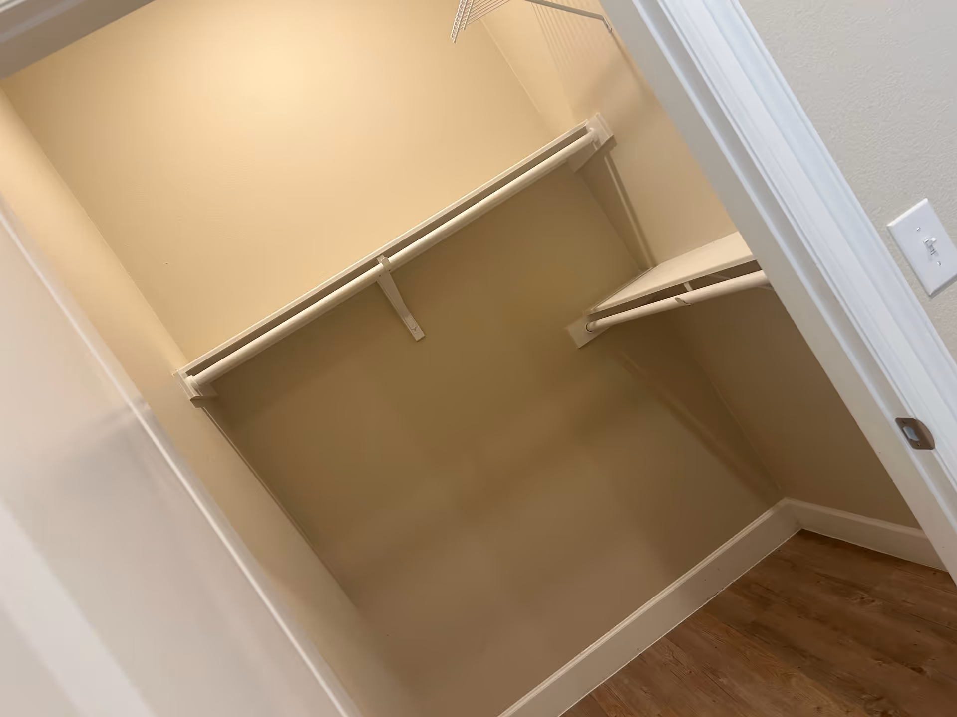 Empty walk-in closet with beige walls, white shelving and hanging rods, and wood flooring.
