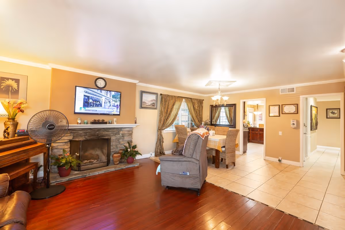 A cozy living and dining area with wooden flooring and tiled floor sections. The living area features a stone fireplace with a mounted TV above it, a standing fan, and potted plants. The dining area has a table with a checkered tablecloth and four wicker chairs, with a chandelier overhead and a large mirror on the wall. The walls are painted beige with framed pictures and certificates, and there is a hallway leading to other rooms.