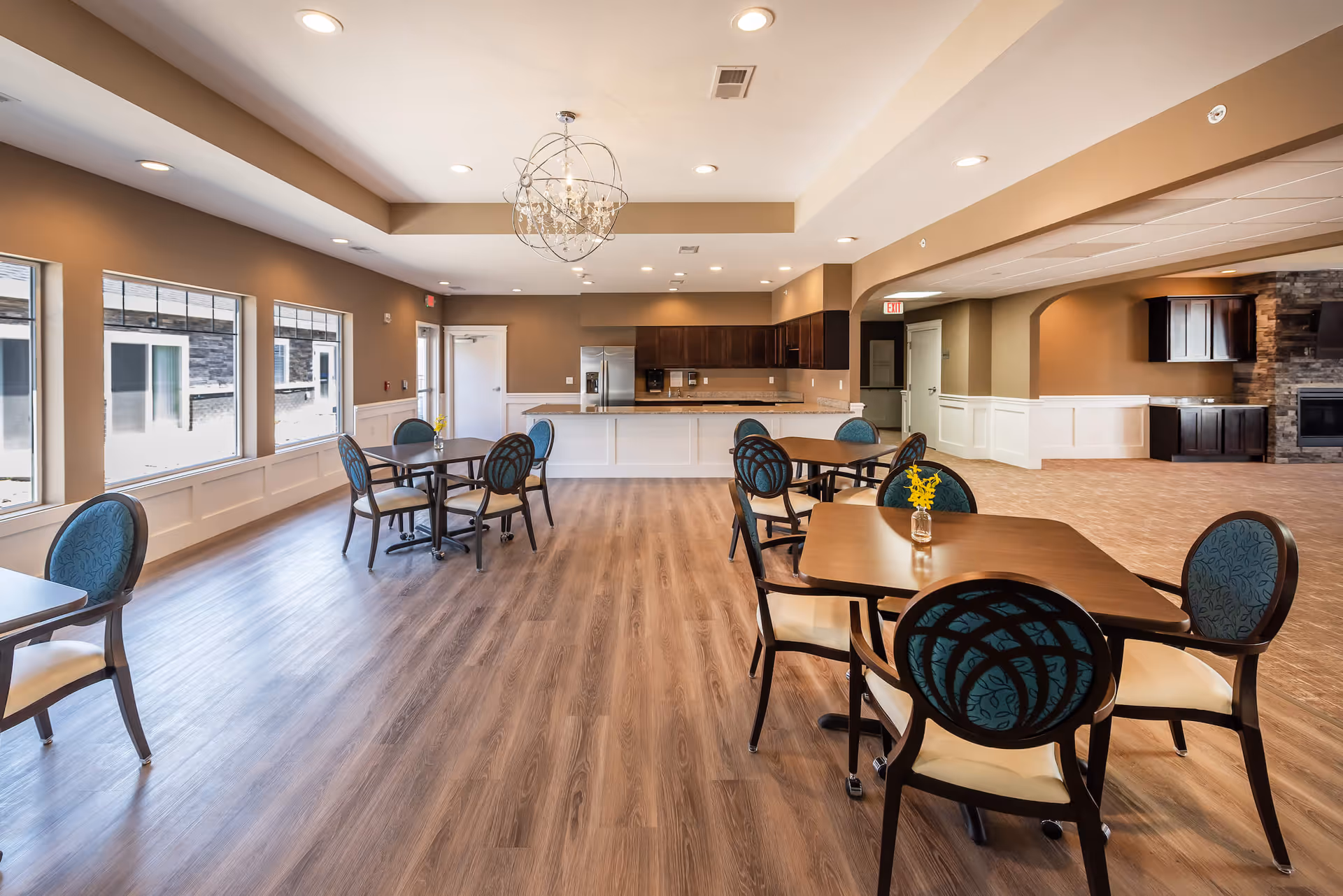 Bright communal dining room with multiple tables and chairs, wood floors, large windows, and a kitchen area in the background.
