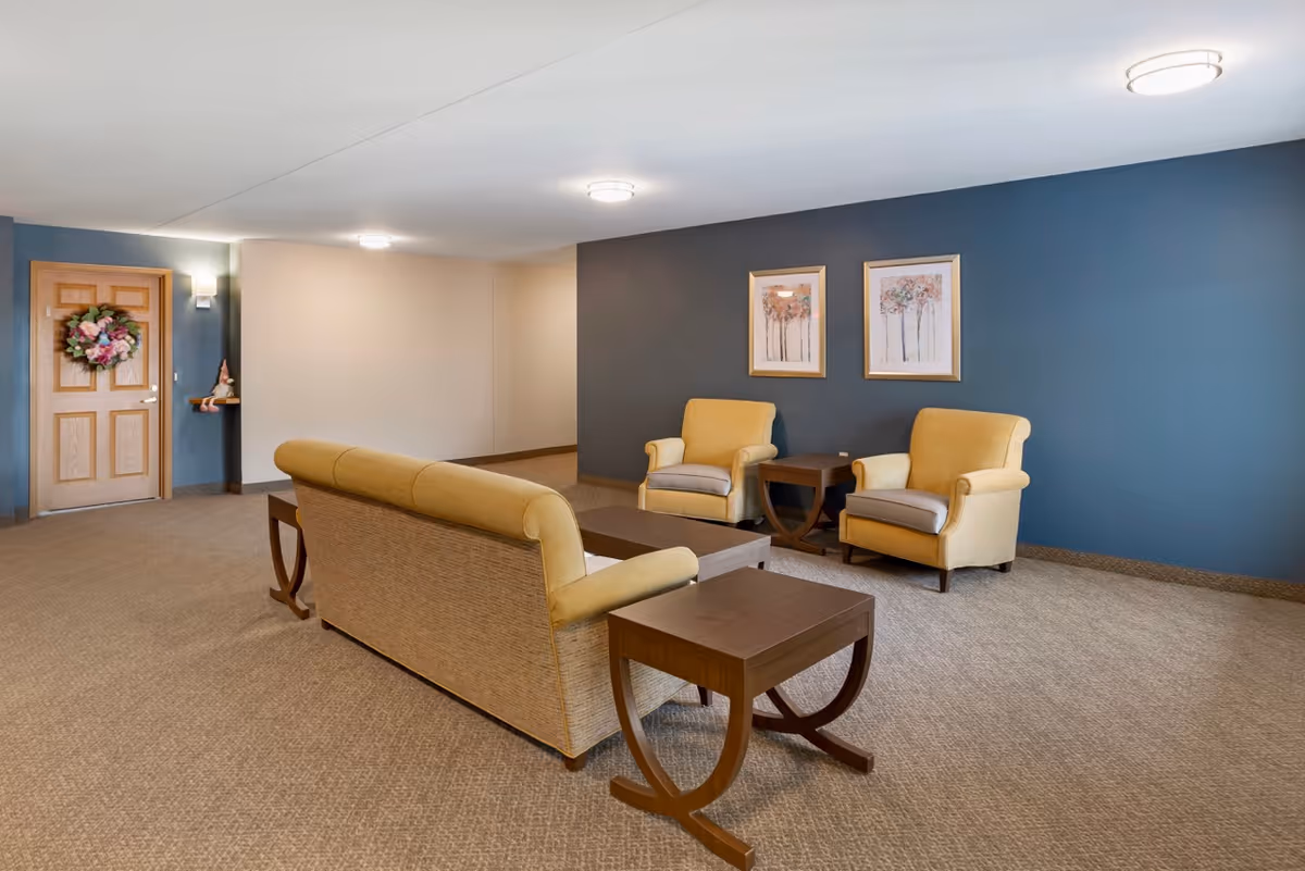 A senior living facility common area with beige carpet, a yellow sofa, two yellow armchairs, and wooden side tables. The walls are painted blue and beige, with two framed floral paintings on the blue wall. A wooden door with a floral wreath is visible on the left side.