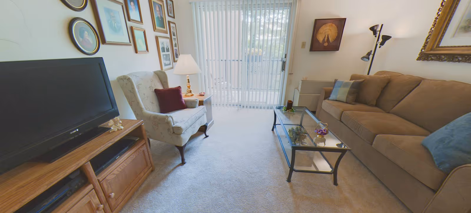 A cozy living room with a brown sofa adorned with blue and green pillows, a glass coffee table with decorative items, a floral armchair with a burgundy pillow, a wooden TV stand with a flat-screen TV, a side table with a lamp, and framed pictures on the wall. Vertical blinds cover a sliding glass door leading outside.