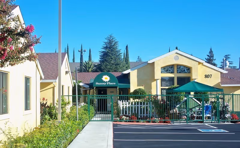 Exterior view of Sunny Place of Stockton, a single-story yellow building with a green awning displaying the facility name. There is a fenced garden area with plants and flowers, a green canopy with chairs underneath, and a parking lot with a handicapped parking space in front. The sky is clear and blue.
