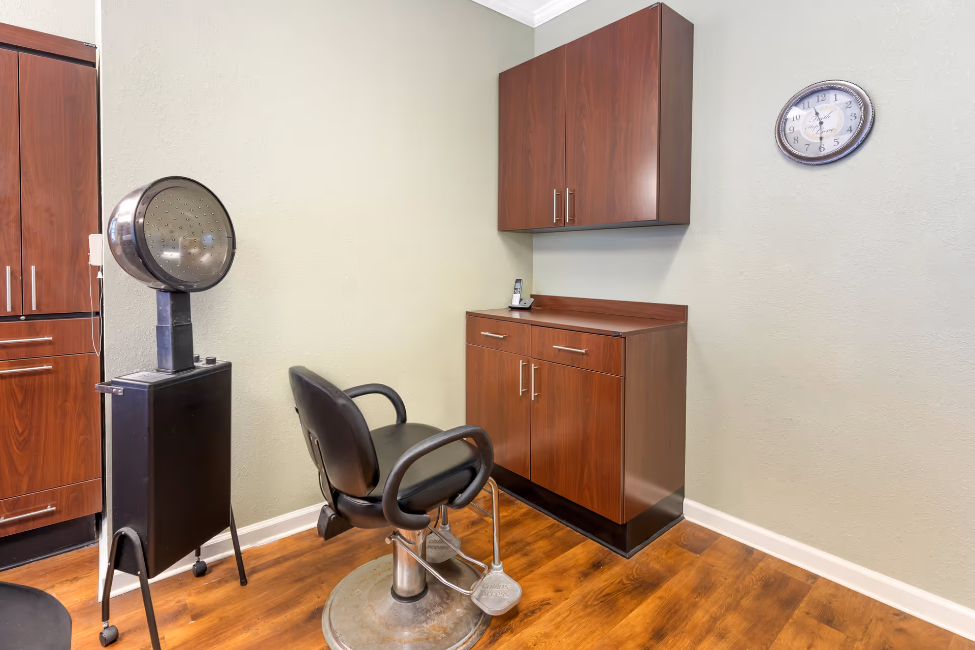 Interior view of a small salon area with a black salon chair, a vintage hair dryer on a stand, wooden cabinets mounted on the wall and below, a cordless phone on the cabinet, and a wall clock showing the time as 12:30.