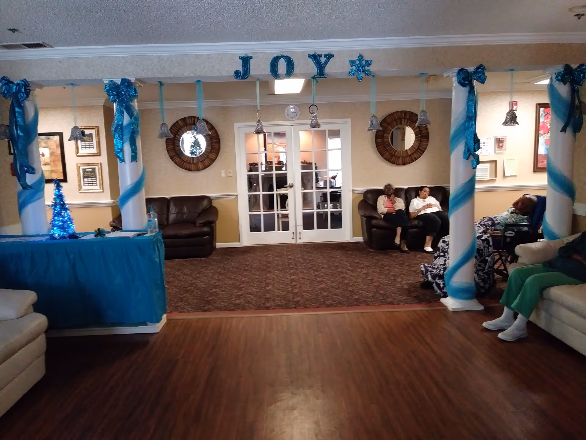 A decorated common lounge with blue ribbons and a 'JOY' sign, columns, couches and several seated residents.