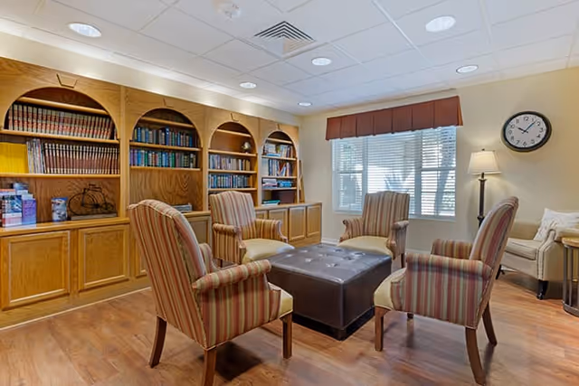 A cozy sitting area in a senior living facility with four striped armchairs arranged around a square brown ottoman. Behind the chairs is a wooden bookshelf filled with books and decorative items. A window with blinds and a valance lets in natural light, and a floor lamp and wall clock are visible on the right side of the room.