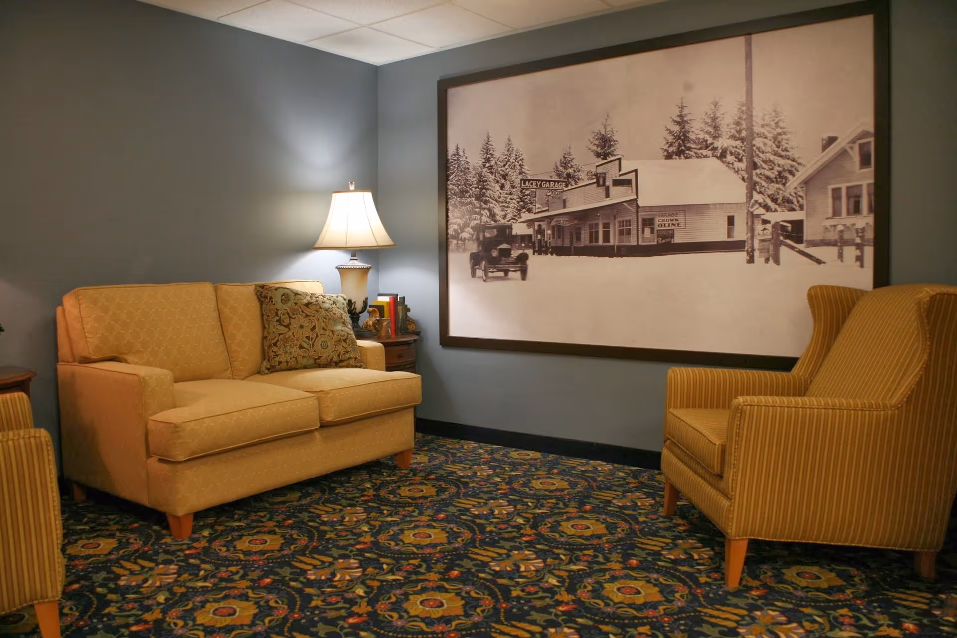 A cozy sitting area with a yellow patterned loveseat and a matching striped armchair on a floral carpet. A table lamp sits on a small wooden side table between the chairs. A large framed black and white photograph of a snowy street scene with buildings and a vintage car hangs on the blue-gray wall behind the seating.