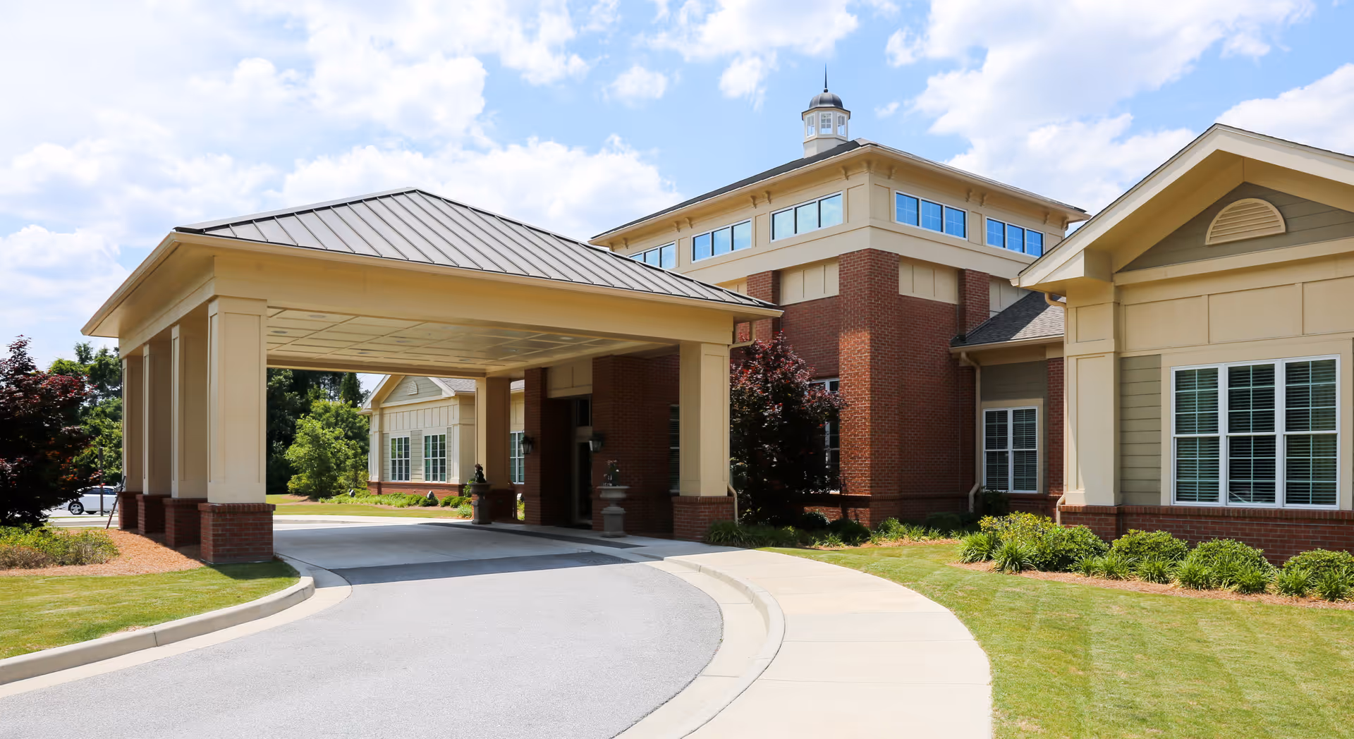 Covered porte-cochère and driveway leading to the brick front of a senior living and rehabilitation building with landscaped lawns and a blue sky.