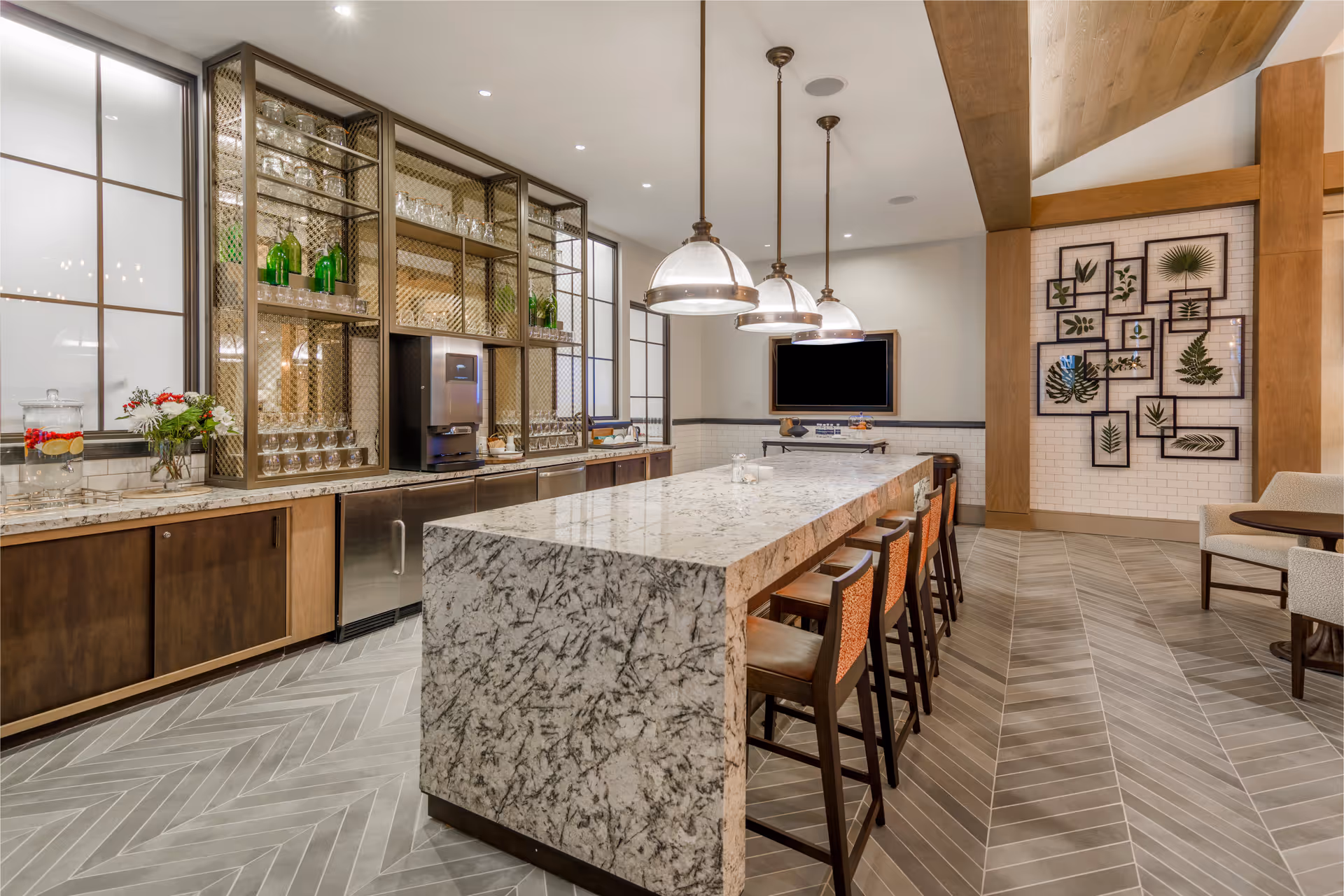 A modern communal kitchen and dining area with a large marble island countertop surrounded by high chairs. The back wall features shelves with glassware and green bottles, a water dispenser, and cabinets below. Three pendant lights hang above the island. The floor has a herringbone tile pattern, and the walls are decorated with framed botanical prints. There is a flat-screen TV mounted on the far wall.