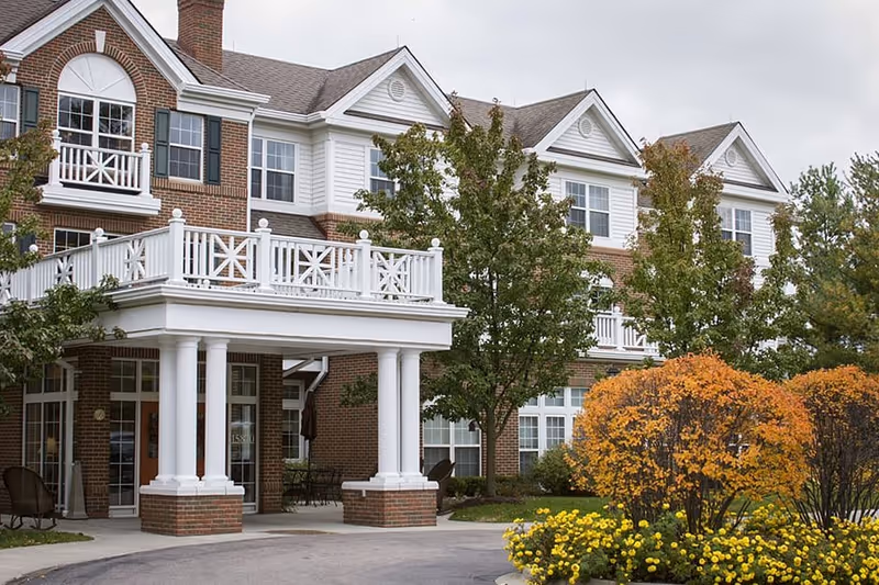 Front entrance of a brick-and-white senior living building with columns, balconies, trees, and landscaped shrubs.
