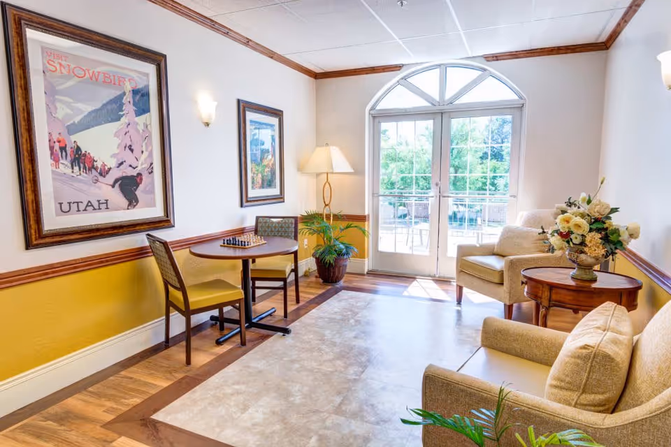 A bright and cozy sitting area in a senior living facility with two beige armchairs, a small round wooden table with a flower arrangement, a round table with two chairs and a chessboard, framed artwork on the walls, a floor lamp, and large glass doors leading outside.