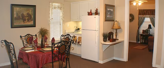Interior view of a dining area with a round table covered with a red tablecloth and set with plates and napkins. The dining area has four wrought iron chairs. Adjacent to the dining area is a small kitchen with white cabinets and a white refrigerator. The walls are beige, and there is a framed painting and a window with blinds. A small shelf with a lamp and decorative items is mounted on the wall near an open doorway leading to a living room with a ceiling fan and additional furniture.
