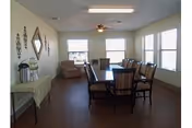 A bright dining room with a rectangular wooden table surrounded by eight chairs. The room has large windows on two walls letting in natural light. There is a ceiling fan with a light fixture above the table. On the left side, there is a small table with decorative items and wall decor including a diamond-shaped mirror and metal wall art.