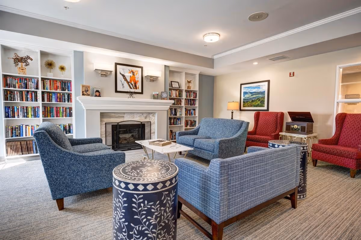 A cozy living room area with a fireplace in the center, flanked by built-in bookshelves filled with books and decorative items. The room features blue upholstered armchairs and a loveseat arranged around a small coffee table, with two red wingback chairs and a vintage-style radio on a side table in the background. The walls are adorned with framed artwork, and the space is well-lit with ceiling lights and a table lamp.