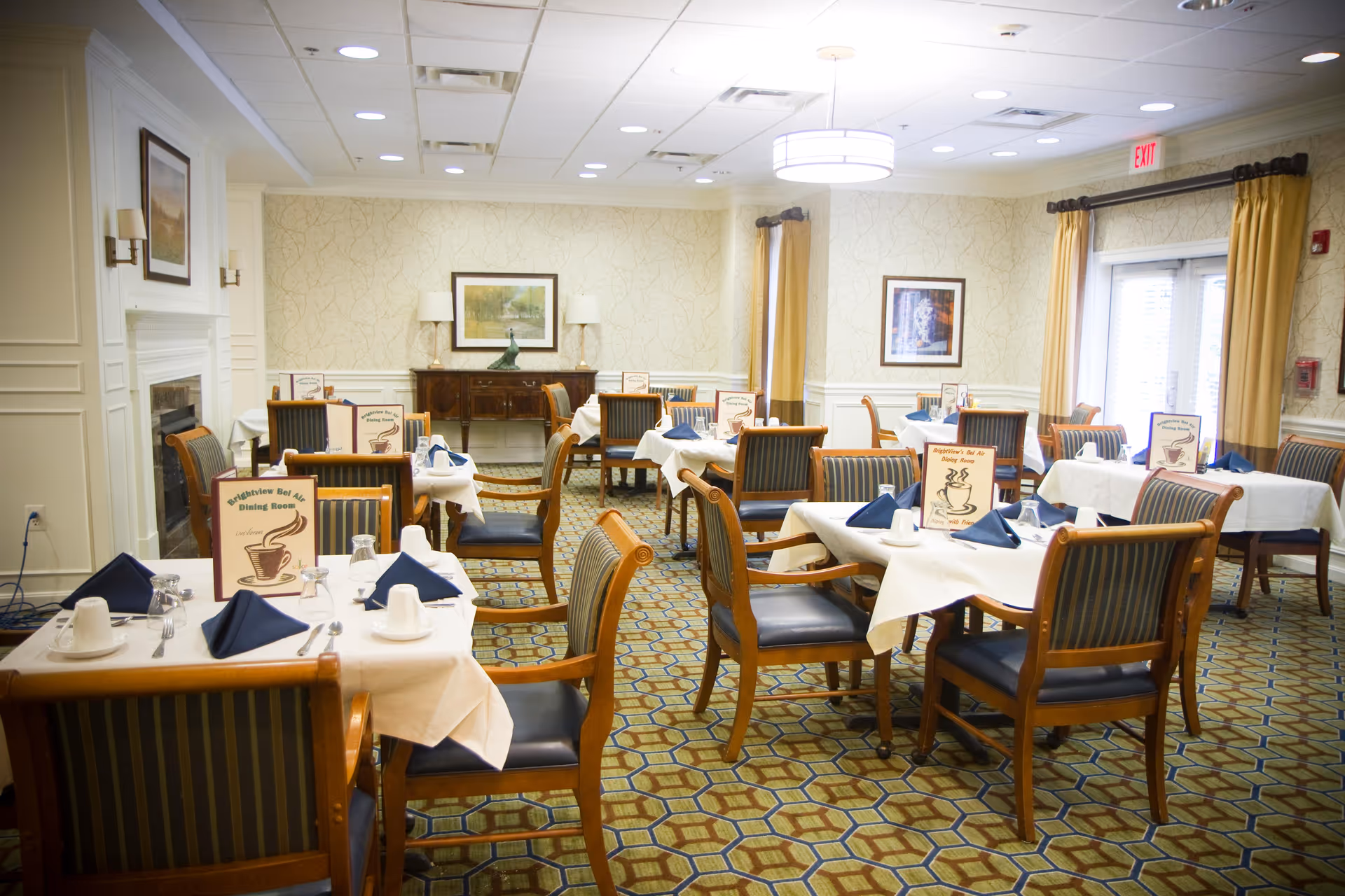 A bright and neatly arranged dining room with multiple tables covered in white tablecloths, each set with blue napkins, cups, glasses, and silverware. The chairs have wooden frames with dark blue striped upholstery. The room features patterned carpet, framed artwork on the walls, yellow curtains, and a fireplace on the left side. Menus labeled 'Brightview Bel Air Dining Room' are placed on each table.