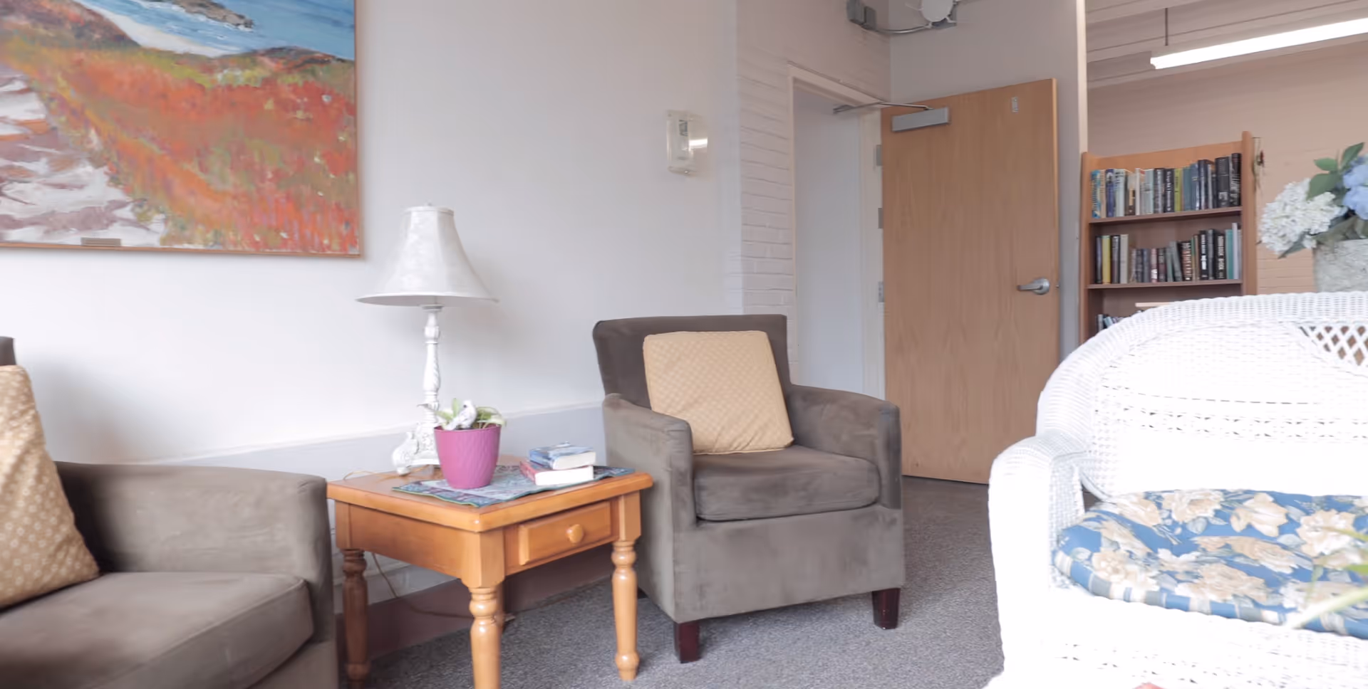 Small lounge with upholstered armchairs, a wooden side table and lamp, a bookshelf and a white wicker chair.