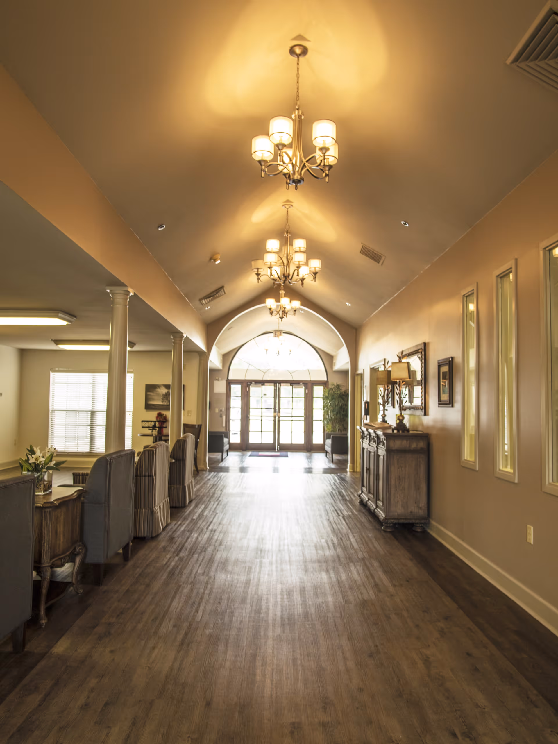 A long, well-lit entrance hallway with chandeliers, wood floors, seating along the sides, and glass double doors at the far end.