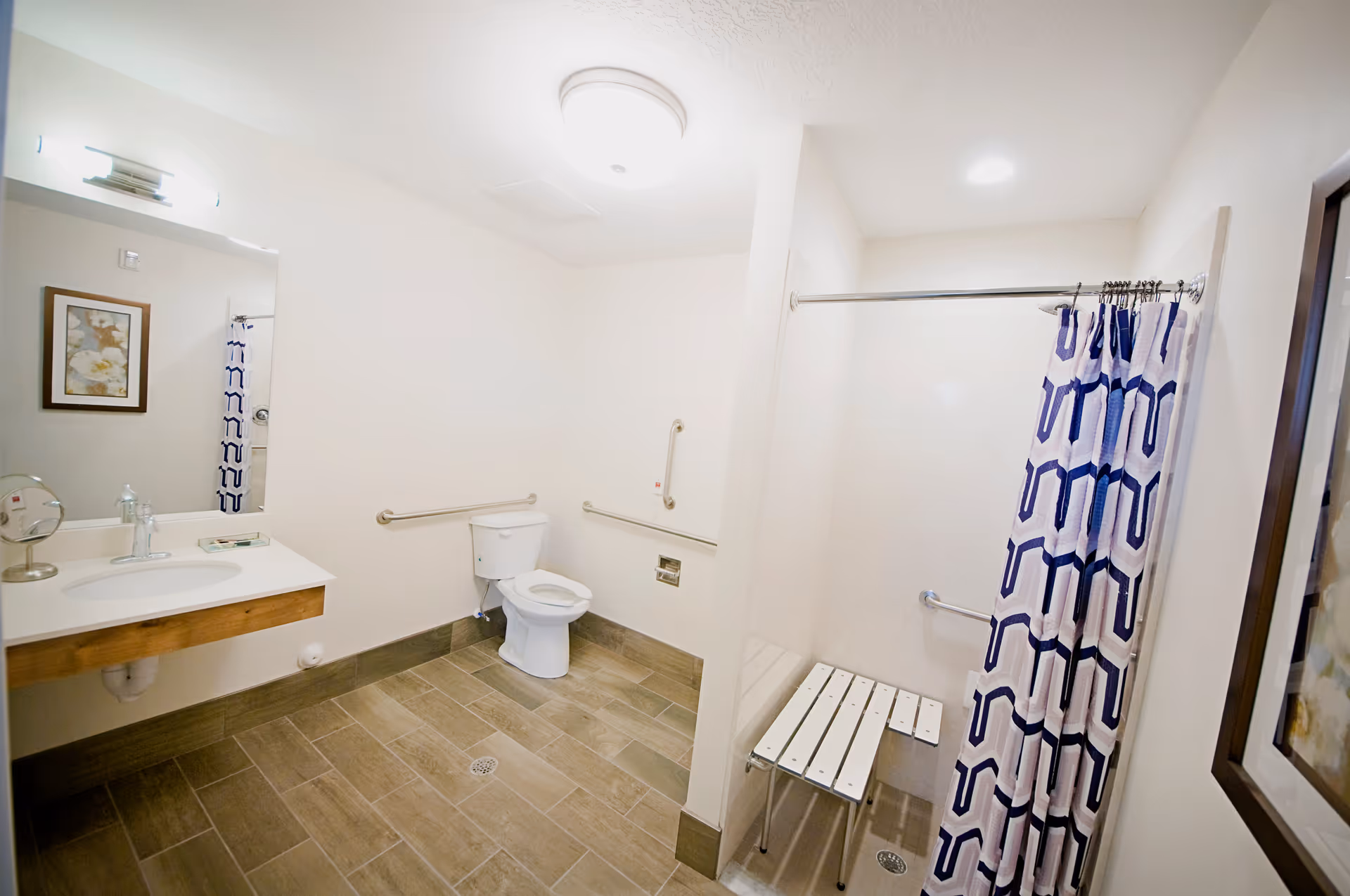 A spacious, accessible bathroom with a toilet, a wall-mounted sink with a mirror above it, grab bars around the toilet, and a walk-in shower with a shower curtain and a fold-down bench seat.