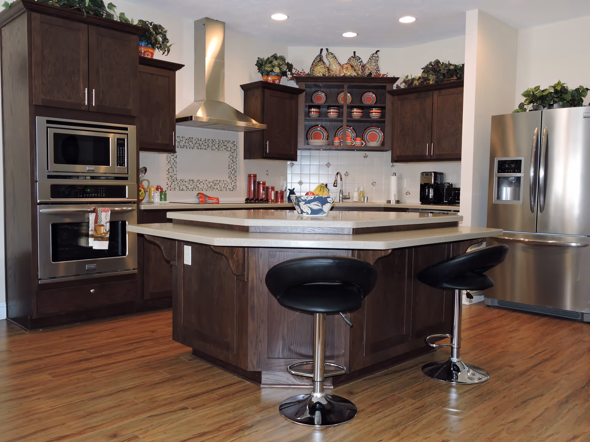 Modern kitchen with dark wood cabinets, stainless steel appliances including a microwave, oven, and refrigerator. There is a central island with a light-colored countertop and two black bar stools. Decorative plates and plants are placed on top of the cabinets and shelves.
