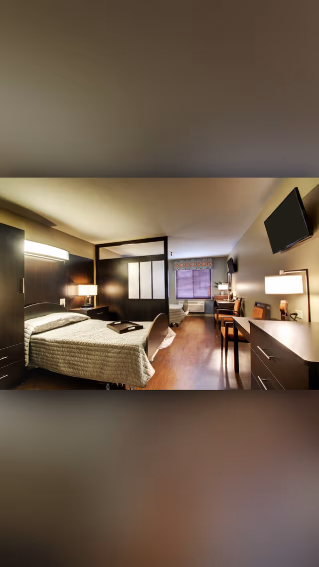 Spacious senior living bedroom suite with an adjustable bed, dark wood furniture, desk, wall-mounted TV, and a window at the far end.