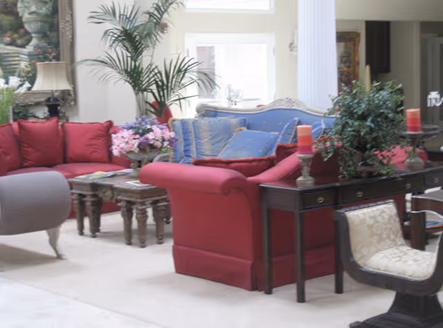 Sunlit living room with red upholstered sofas, decorative pillows, side tables with candles and plants, and a coffee table with flowers.
