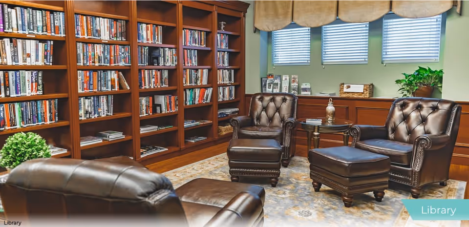 A cozy library room with wooden bookshelves filled with books, three dark brown leather armchairs with matching ottomans, a small round side table, a patterned rug, and windows with blinds and valances. There are also some plants and brochures on a wooden cabinet under the windows.
