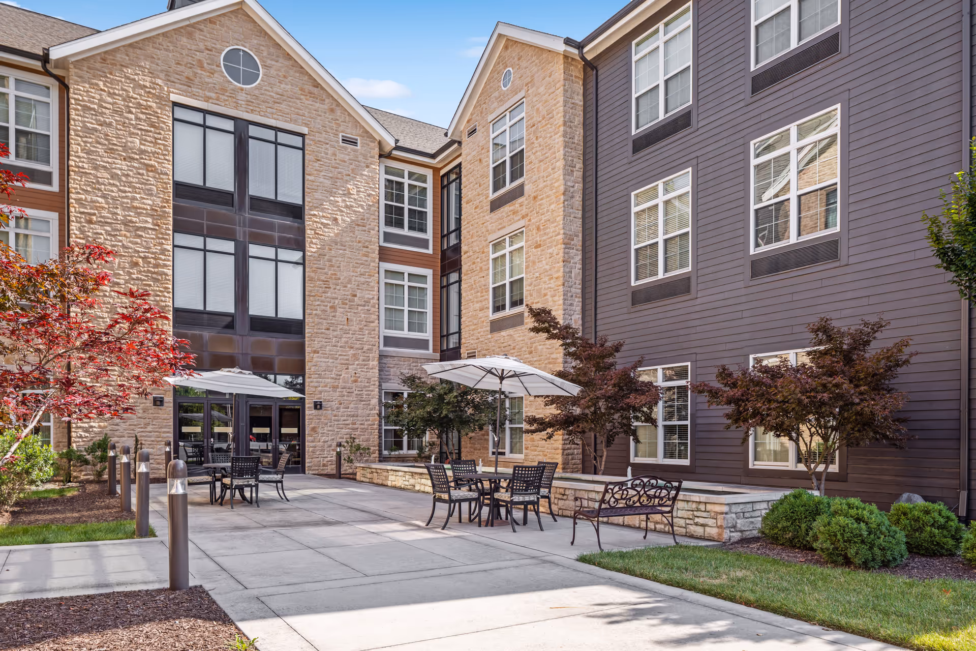Outdoor courtyard area of a senior living facility with patio tables, chairs, and umbrellas. The building has a combination of stone and siding exterior with multiple windows. There are small trees and shrubs planted around the paved seating area.