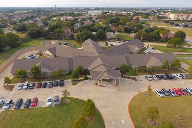 Aerial view of Legend Oaks Healthcare and Rehabilitation - North Austin, showing a large single-story building with a complex roof structure surrounded by parking lots with numerous cars. The facility is situated in a suburban area with many houses and trees visible in the background.