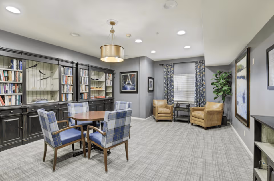 A cozy interior room with a round wooden table surrounded by four blue plaid upholstered chairs. Behind the table is a large black bookshelf filled with books and decorative items. The room has gray walls, a patterned carpet, two tan leather armchairs near a window with floral curtains, and framed artwork on the walls. A modern pendant light hangs from the ceiling.