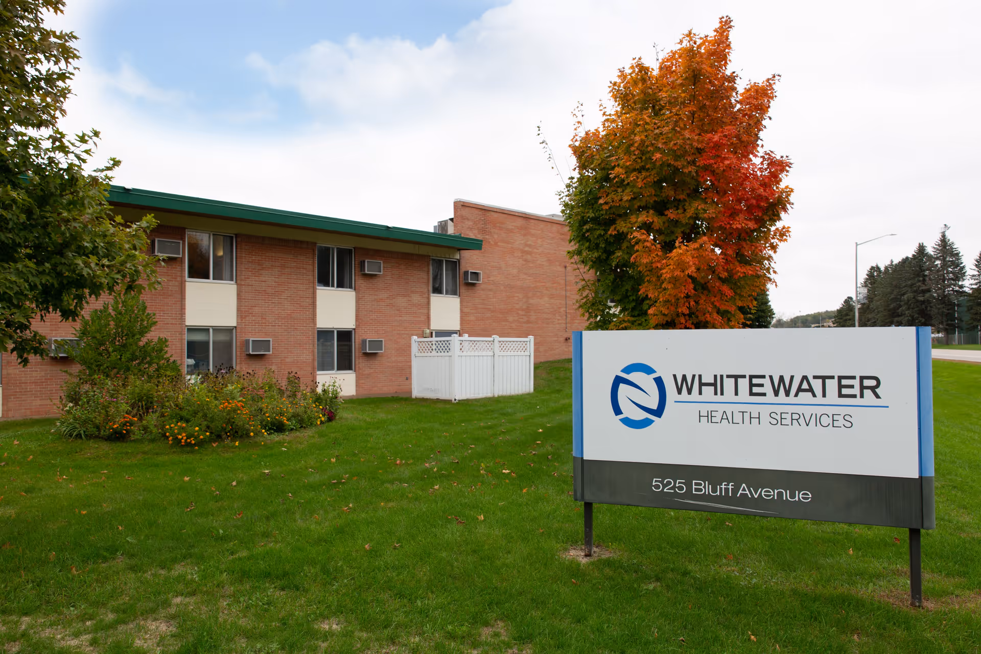 Exterior view of Whitewater Health Services building with a green lawn, trees with autumn foliage, and a sign displaying the facility name and address 525 Bluff Avenue.