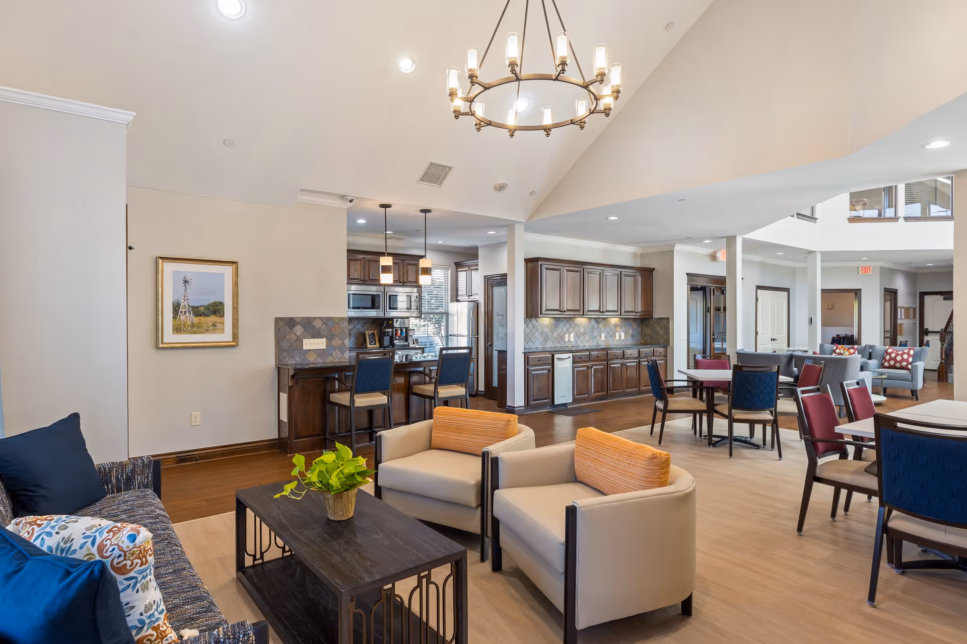A spacious and well-lit common area in a senior living facility featuring a living room with a sofa, two armchairs with orange cushions, and a dark wooden coffee table with a small green plant. In the background, there is a kitchen area with dark wooden cabinets, a tiled backsplash, and a breakfast bar with two chairs. Several dining tables and chairs are arranged throughout the space, and a large chandelier hangs from the high ceiling.