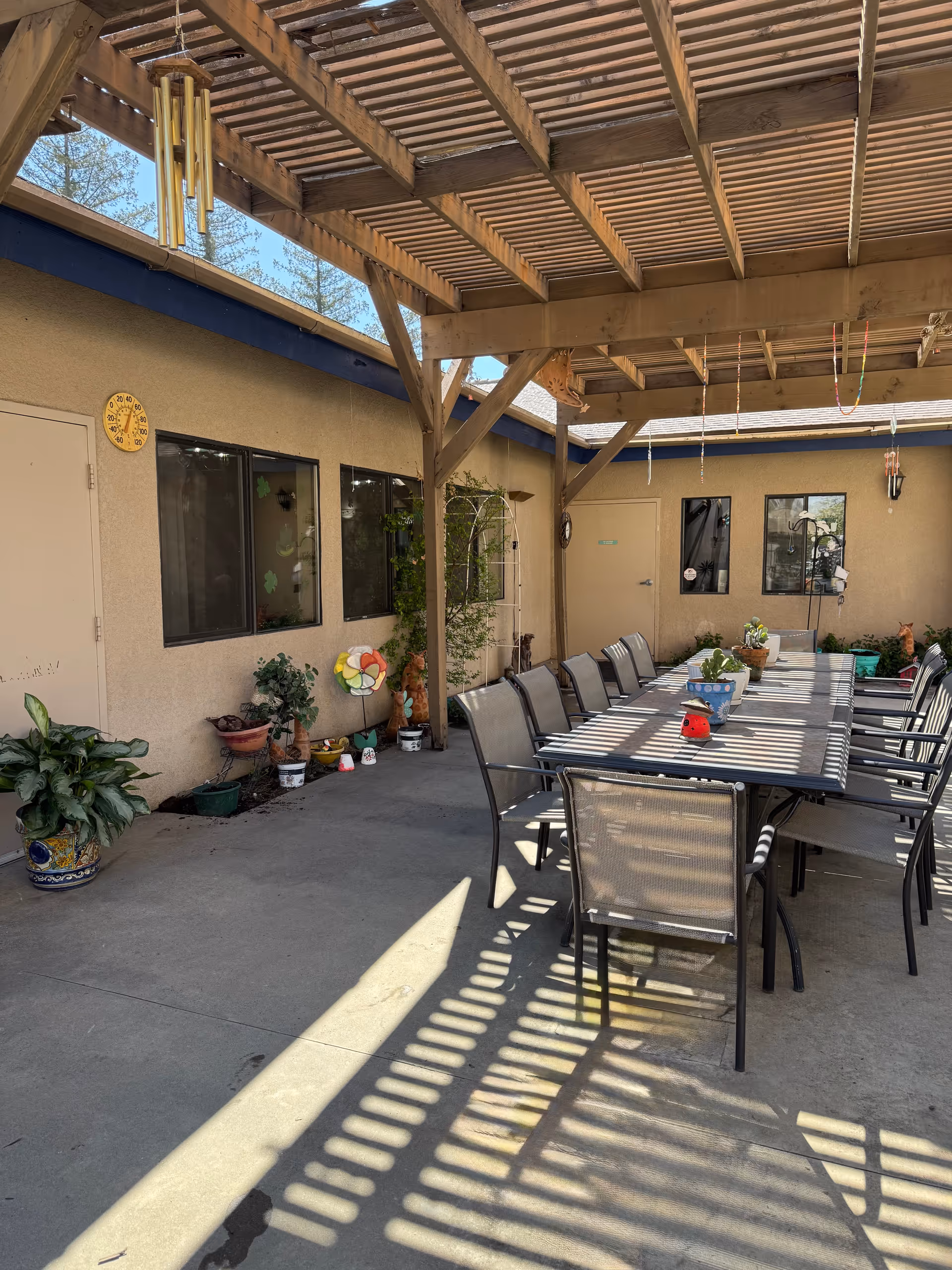 Covered outdoor courtyard with a long dining table and many chairs under a wooden pergola and potted plants along the wall.