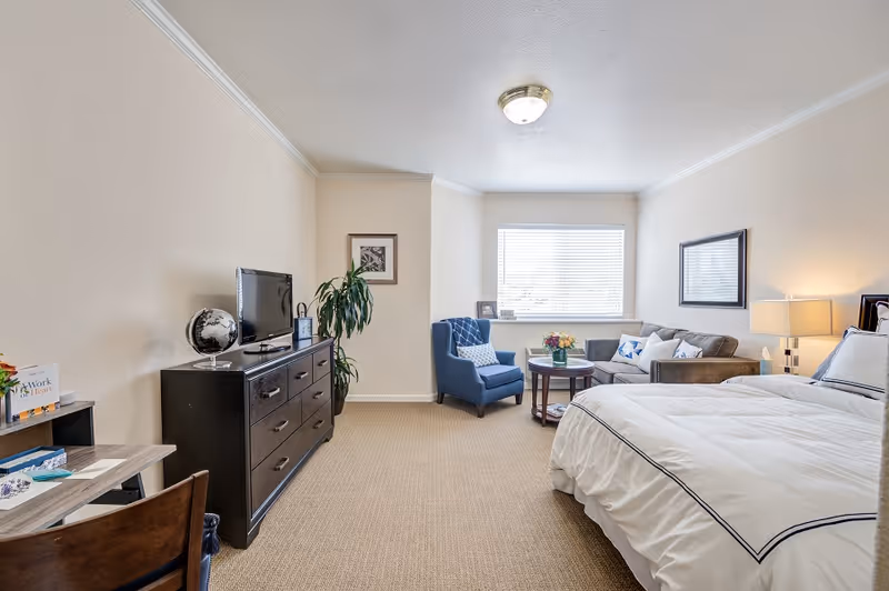 Bright furnished studio bedroom with a bed, dresser and TV, and a seating area by a window.