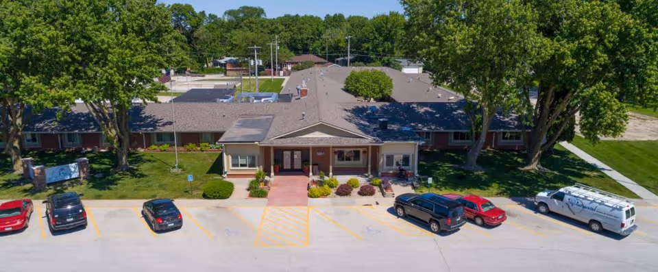 Aerial view of a single-story health and rehabilitation facility building with a front entrance, surrounded by green trees and a parking lot with several cars parked. The building has a brown roof and a well-maintained lawn with shrubs and flowers near the entrance.