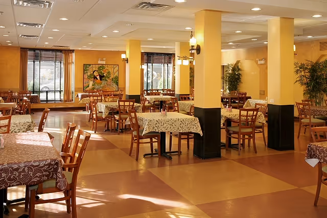 A spacious dining room with multiple wooden tables and chairs. The tables are covered with patterned tablecloths in various colors. The room has yellow walls with black accents on the lower part of the columns, large windows with blinds, and wall-mounted light fixtures. There are plants and a colorful painting on the far wall.