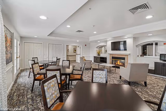 A spacious common area in a senior living facility featuring multiple tables and chairs with patterned upholstery, a fireplace with a flat-screen TV mounted above it, and comfortable armchairs arranged around a coffee table. The room has a neutral color palette with white walls, recessed lighting, and carpeted flooring. There are doors and windows along the walls, and decorative artwork is visible.