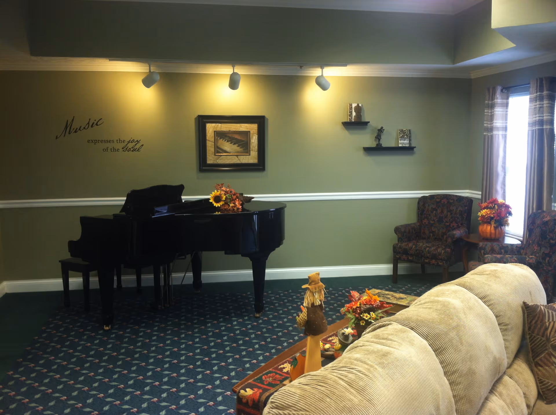 A cozy living room area with a black grand piano adorned with a small flower arrangement. The wall behind the piano has a framed picture of piano keys and a decorative quote that reads 'Music expresses the joy of the soul.' Two patterned armchairs and a small table with a flower arrangement are near a window with curtains. A beige couch is partially visible in the foreground with a decorative centerpiece on the coffee table.