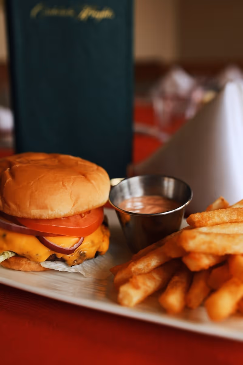 Close-up of a cheeseburger with tomato and onion, a metal cup of sauce, and a side of French fries on a white plate.
