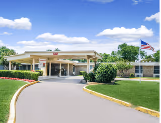Front exterior view of Desoto Long Term Care Partners building with a covered entrance, a curved driveway, well-maintained green lawns, bushes, flower beds, and an American flag on a flagpole under a partly cloudy blue sky.