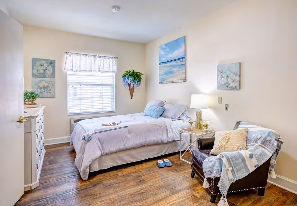A cozy bedroom with a neatly made bed covered in light purple bedding, a pair of blue slippers on the wooden floor, a white dresser with plants on top, a window with blinds and a lace curtain, a side table with a lamp and a glass, a comfortable armchair draped with a blue and white blanket and a pillow, and floral and beach-themed wall art.