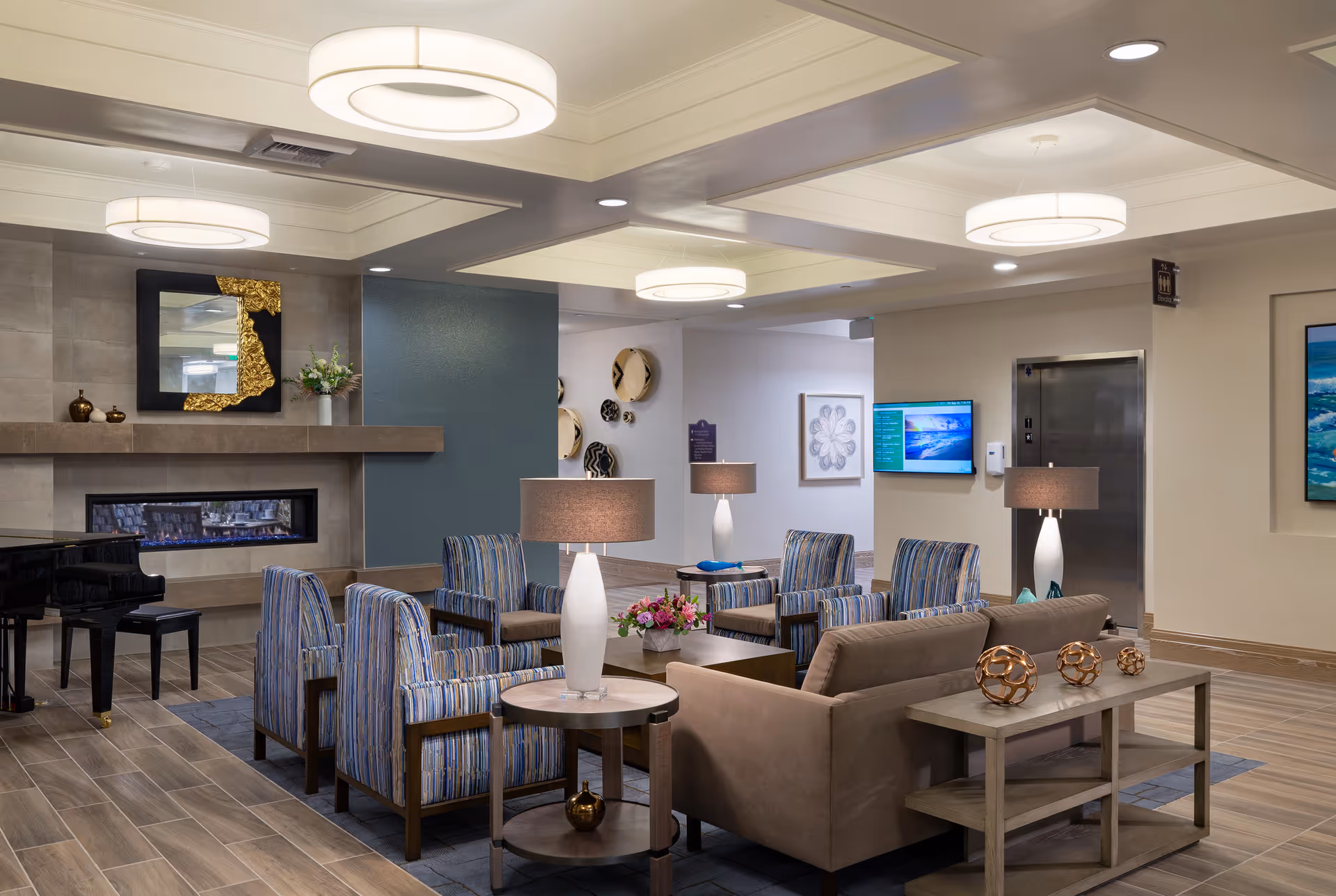 A modern and comfortable living room area in a senior living facility featuring a beige sofa, four striped armchairs arranged around a coffee table with a flower arrangement, two side tables with white lamps, a decorative fireplace with a mirror above it, a black grand piano, and an elevator in the background. The room has wood-look tile flooring, recessed lighting, and decorative wall art.
