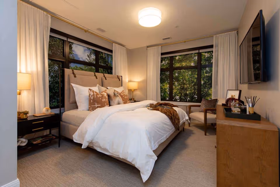 Spacious bedroom featuring a neatly made bed with decorative pillows, bedside lamps, large windows with curtains, an armchair, and a wall-mounted TV.