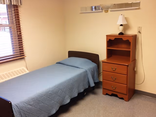 A simple bedroom with a single bed covered with a blue quilted bedspread, a wooden nightstand with three drawers and an open shelf, and a small lamp on top. There is a window with wooden blinds on the left wall and a wall-mounted light fixture above the bed.