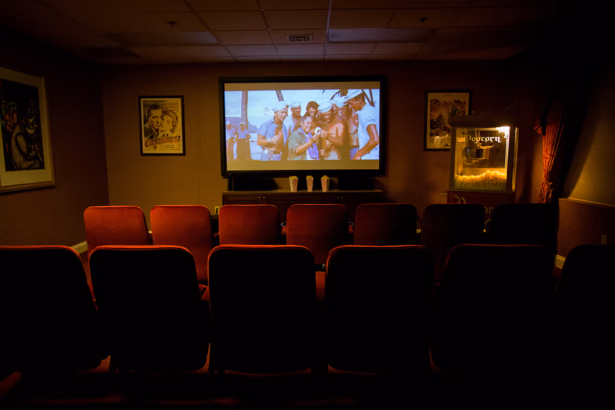 A small movie theater room with two rows of red cushioned seats facing a large screen showing a classic movie scene with sailors. There are three popcorn containers on a cabinet below the screen and a popcorn machine illuminated on the right side of the room. The walls are decorated with framed movie posters.